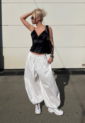 Mujer rubia con top negro sin mangas, pantalones blancos holgados, zapatillas deportivas blancas y gafas de sol, de pie contra una pared clara, con la mano en el cabello.