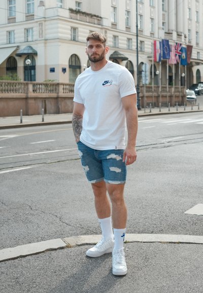 Camiseta blanca de Nike con logo, pantalones cortos de mezclilla desgastados y zapatillas blancas. Los calcetines tienen un diseño azul. Entorno urbano con edificios de fondo.