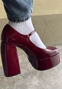 Bordojas laku ādas platformas kurpes ar apaļu purngalu, biezu blokpapēdi un potītes siksniņu, kopā ar baltu zeķi.