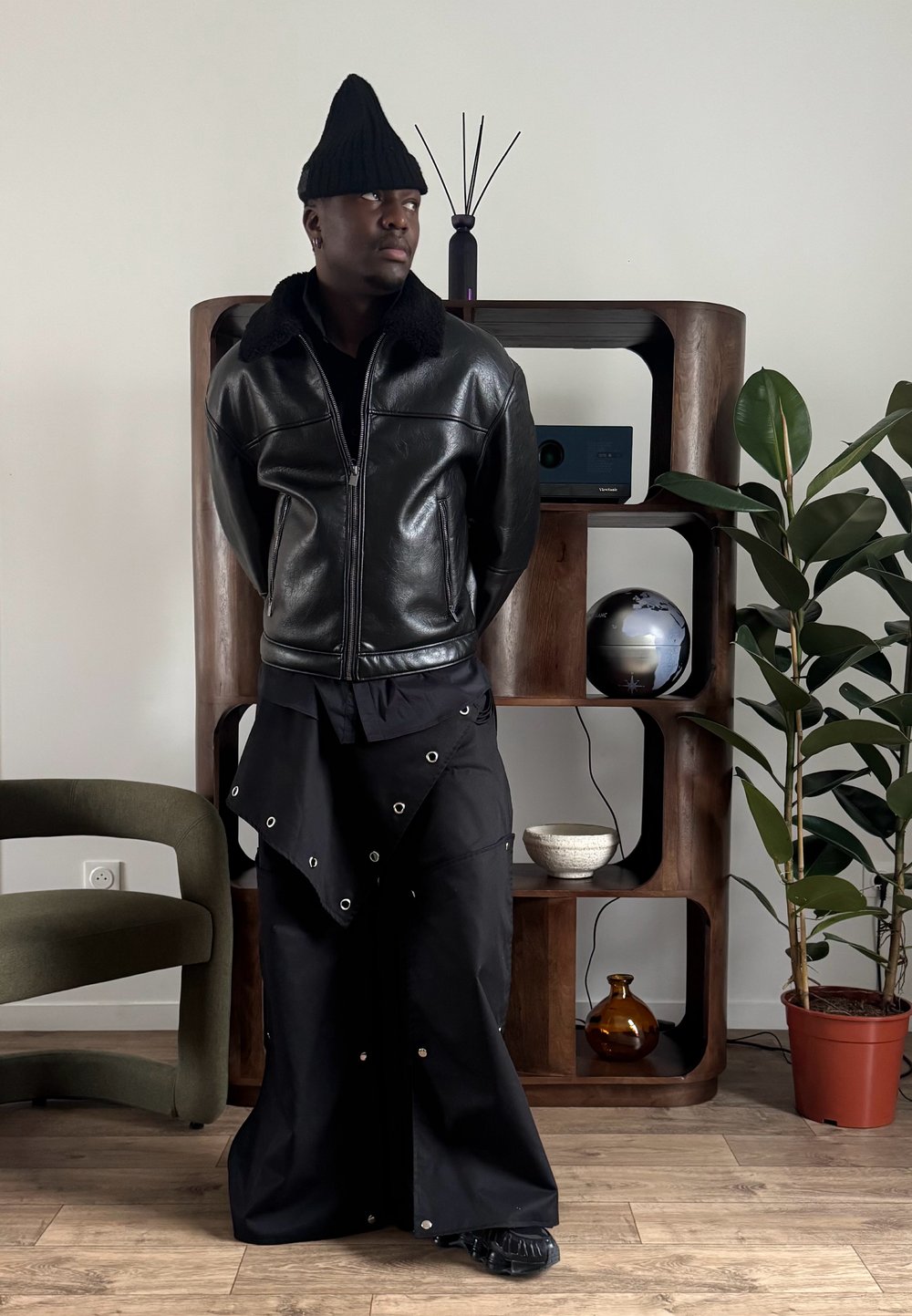 Veste en cuir noire avec col en fourrure, pantalon noir ample avec œillets argentés, et un bonnet en tricot noir à pointe. Cadre intérieur avec des plantes.