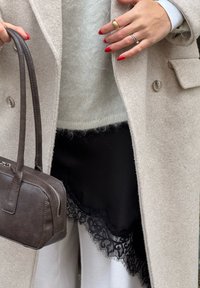 Femme portant un manteau beige sur un pull blanc duveteux et une jupe noire avec bordure en dentelle, tenant un petit sac à main en cuir brun foncé.