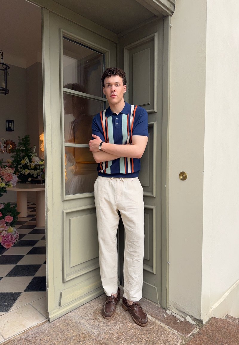 Young man with curly hair stands with arms crossed in a doorway, wearing a striped navy shirt, white trousers, and brown shoes near flowers inside.