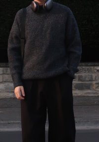 Mørkegrå strikket sweater med et tekstureret mønster, parret med sorte brede bukser. Høretelefoner hviler om halsen, hænderne i lommerne.