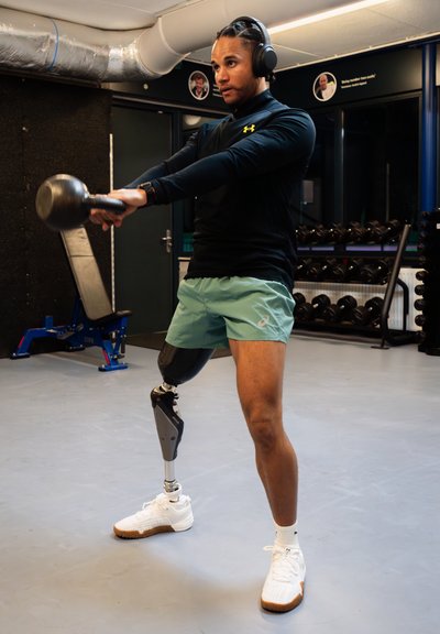 Atleta con camiseta de manga larga negra y pantalones cortos verde menta realiza un movimiento de kettlebell swing. Tiene una pierna prostética izquierda y zapatillas deportivas blancas.