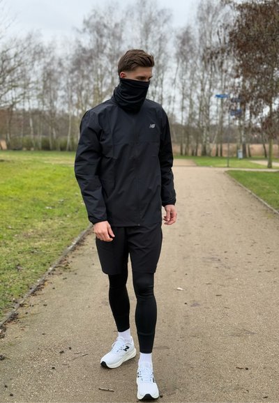 Chaqueta ligera negra, bufanda de cuello, pantalones cortos negros y leggings negros. Zapatillas deportivas blancas con suelas texturizadas. Entorno de parque al aire libre.