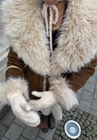 Personne portant un manteau marron avec un large col et des poignets en fourrure claire, des moufles tricotées crème, debout sur un trottoir en pavés près d'une lumière circulaire au sol.