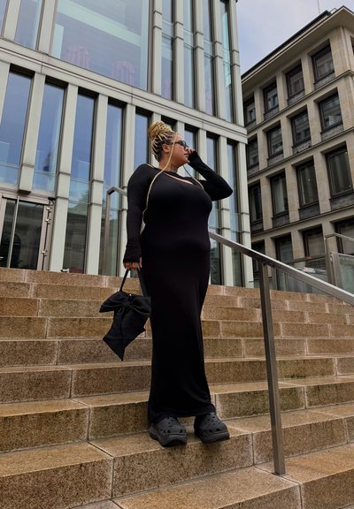 Vestido negro de manga larga con escote recortado, crocs negros y un bolso negro con diseño de lazo, en un entorno de arquitectura moderna de vidrio.