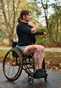 Černá sportovní bunda a zelené šortky na osobě na invalidním vozíku. Detailní květinové tetování na noze. Zlaté akcenty na kolech invalidního vozíku.