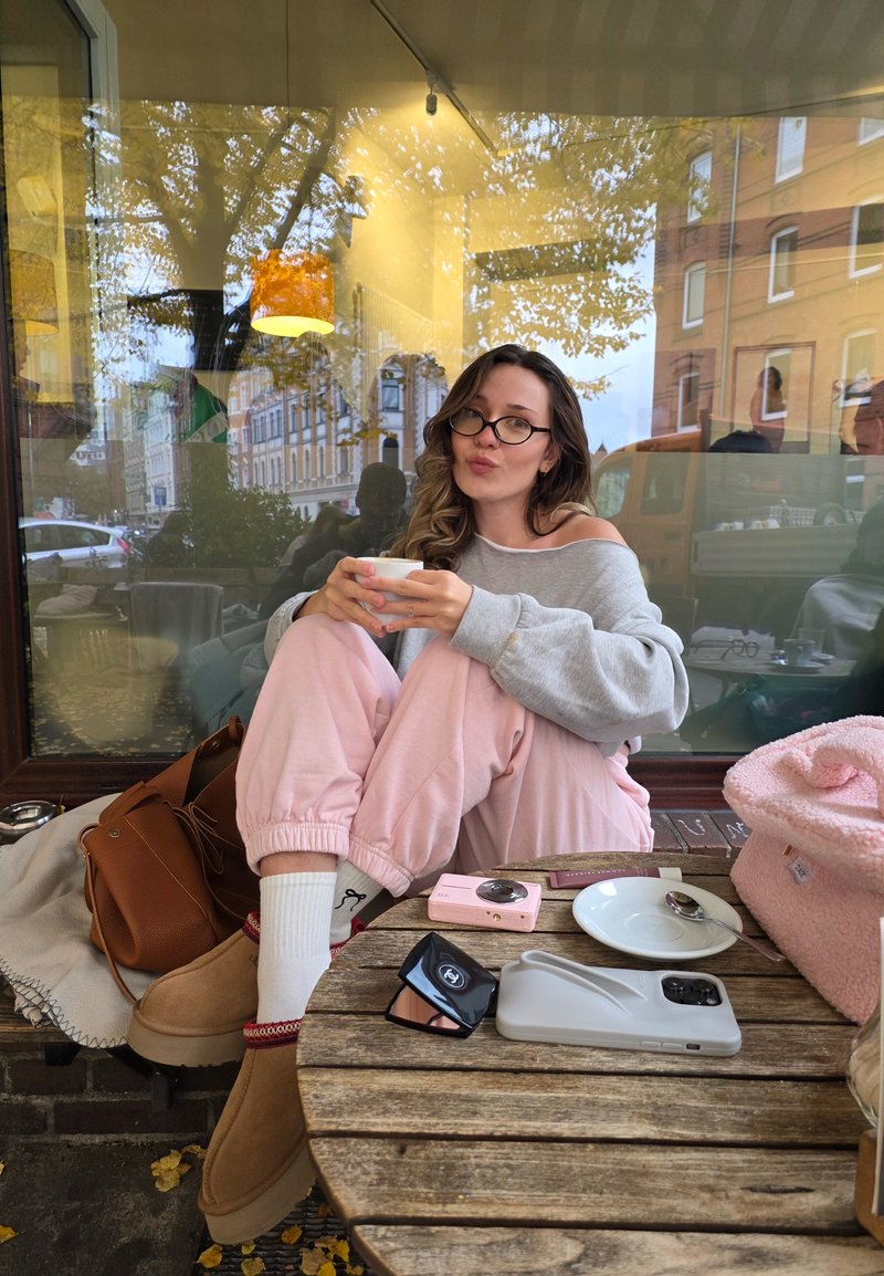 Person in gray oversized sweater and pink sweatpants sitting on a wooden table outdoors, holding a white cup, with accessories and a pink bag nearby.