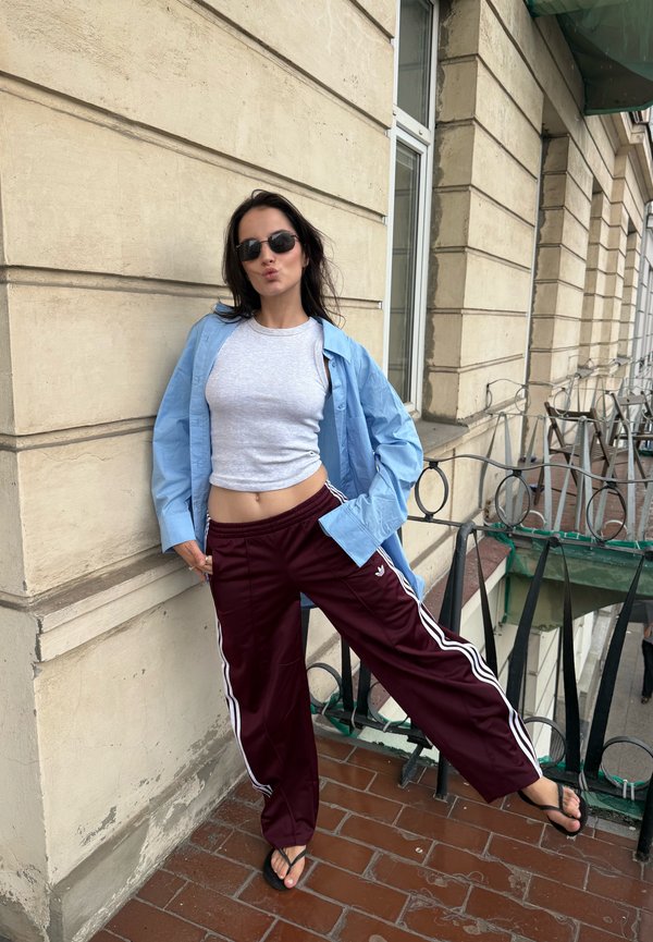 Femme portant des lunettes de soleil, une chemise bleue, un crop top gris et un pantalon de survêtement bordeaux, appuyée contre un mur beige d'un bâtiment sur un balcon carrelé.