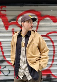 Μπέζ φούτερ με φερμουάρ πάνω από μαύρο πουκάμισο με κουμπιά και μπεζ ριγέ μπλούζα baseball, με καφέ καπέλο και αξεσουάρ σκουλαρίκι. Γκρίζα τοιχογραφία με κόκκινα γκράφιτι.