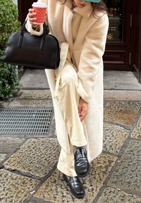 Fehér plüss hosszú kabát, bézs kordbársony nadrág, fekete bőr táska két fogantyúval, és fekete loaferek csattal díszítve. Egy kávéscsészét tart a kezében.