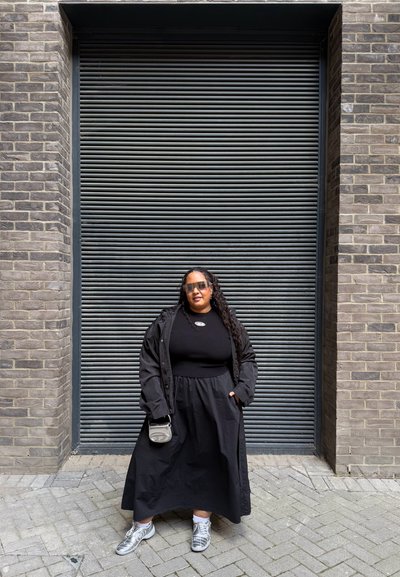 Abrigo negro de gran tamaño sobre un top negro acanalado y una falda negra fluida. Llevado con zapatillas plateadas y gafas de sol, sosteniendo un pequeño bolso gris.