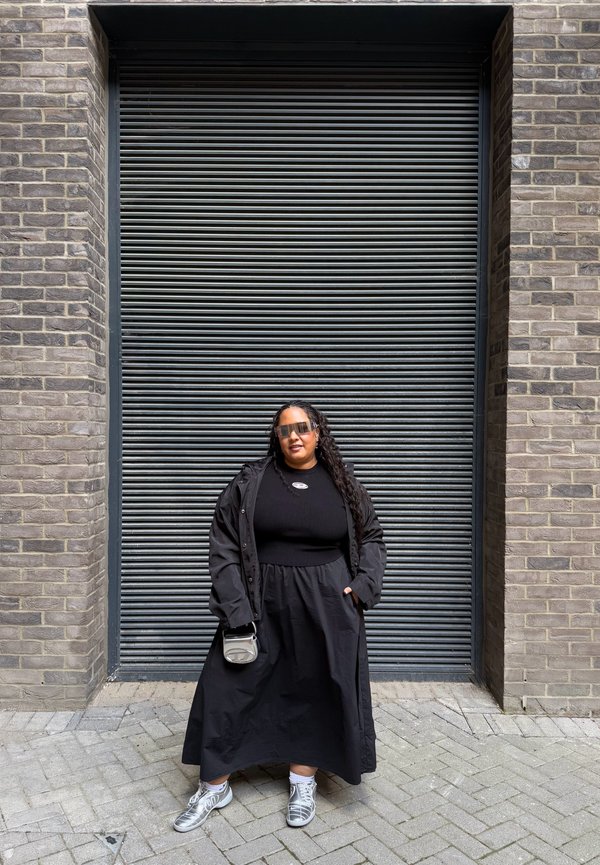 Oversized black coat over a black ribbed top and a flowing black skirt. Paired with silver sneakers and sunglasses, carrying a small grey bag.