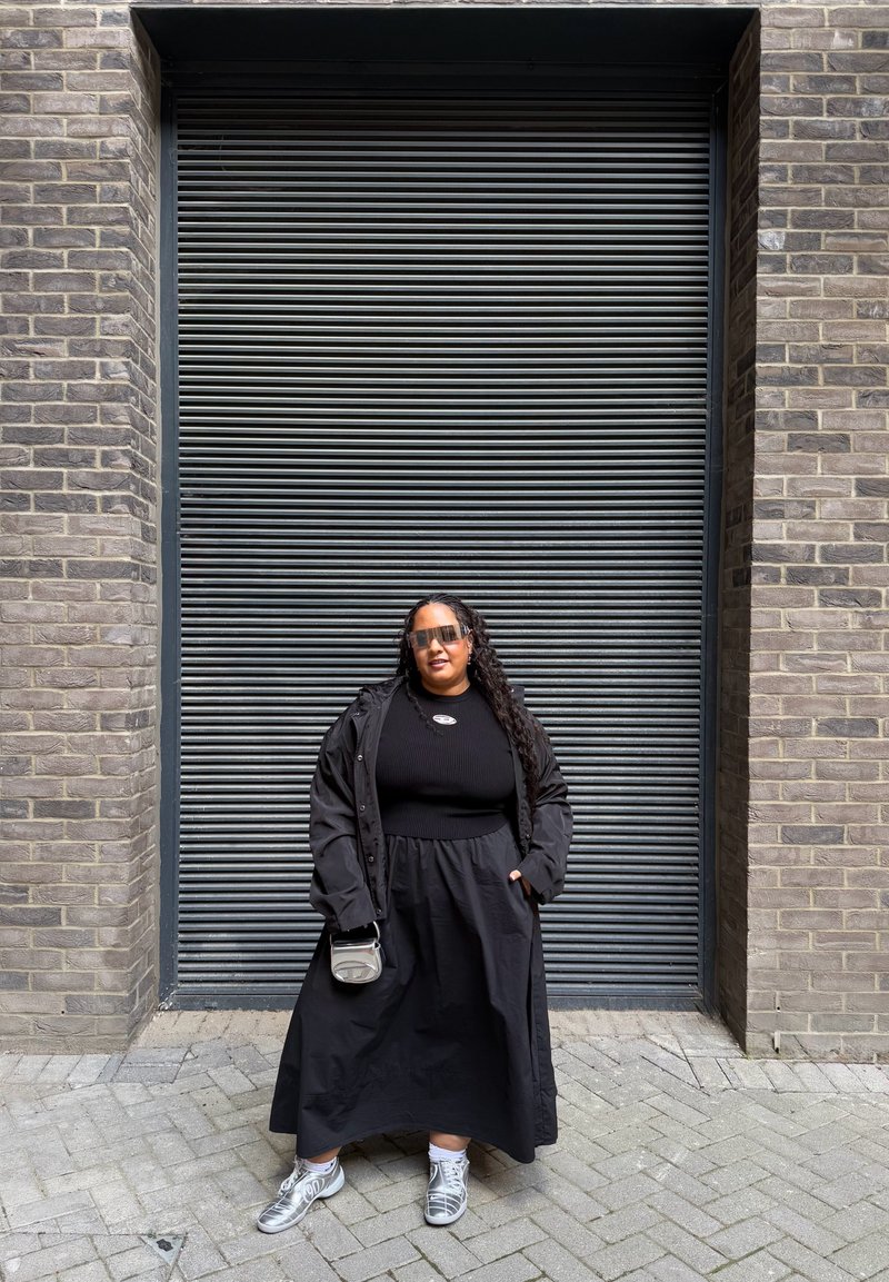Casaco oversized preto sobre um top preto com nervuras e uma saia preta fluida. Usado com tênis prateados e óculos de sol, segurando uma pequena bolsa cinza.