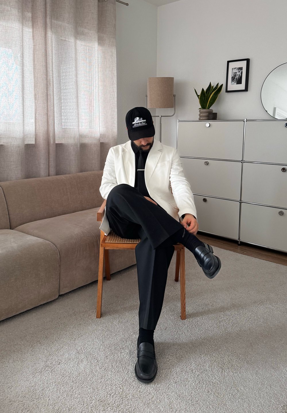 Homme en pantalon noir, chaussures noires, veste blanche et casquette noire, assis les jambes croisées dans un salon moderne avec canapé et meuble.