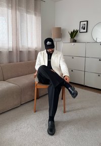 Homme en pantalon noir, chaussures noires, veste blanche et casquette noire, assis les jambes croisées dans un salon moderne avec canapé et meuble.