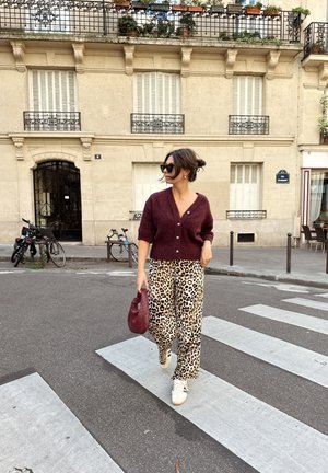Cardigan de punto en color burdeos, pantalones de pierna ancha con estampado de leopardo, zapatillas blancas y un bolso rojo. Mujer cruzando una calle con un edificio decorativo al fondo.