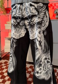 Pantalon noir avec des motifs blancs complexes d'ailes, d'étoiles et de volutes, porté avec un pull gris à l'intérieur.