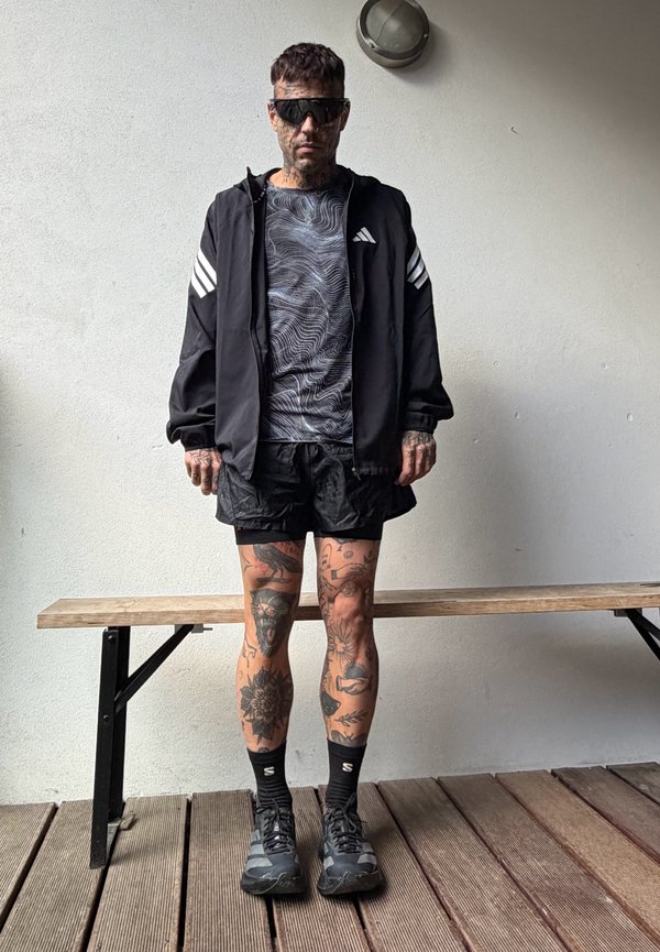 Un hombre tatuado con gafas de sol, una chaqueta deportiva negra, una camisa estampada, pantalones cortos y zapatillas está de pie sobre una terraza de madera.