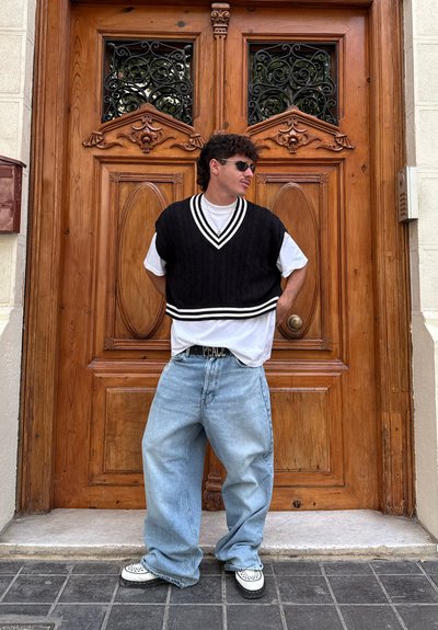 Suéter negro recortado con rayas blancas sobre una camiseta blanca, jeans claros y holgados, y zapatillas en blanco y negro, posando frente a una puerta de madera.