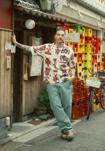 Camisa de manga corta con base crema y patrones gráficos en rojo y negro. Pantalones verde holgados y zapatos de estampado de leopardo. Entorno urbano.