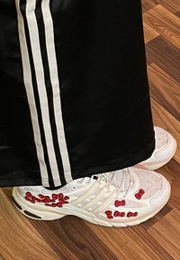 Bele tene najke z rdečimi poudarki Hello Kitty, ki imajo teksturirane vzorce in oblazinjen podplat, oblečene z črnimi hlačami z belimi stranskimi črtami.