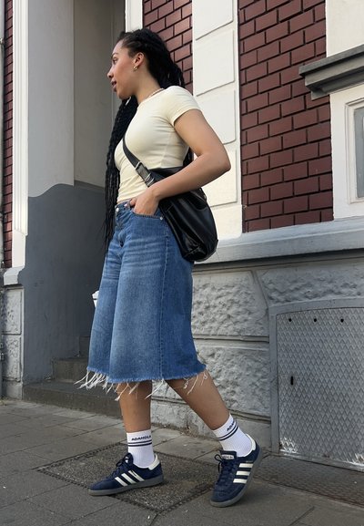 Falda de mezclilla con dobladillo deshilachado, top ajustado de color amarillo claro, bolso negro y zapatillas oscuras con rayas blancas y calcetines estampados, de pie sobre el pavimento.