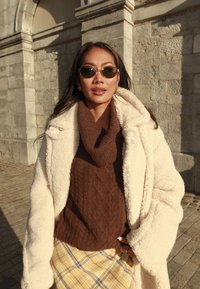 Kreemjas sametmantel pruunile kootud kampsuni, mustrilise kollase ruudulise seeliku ja ümmarguste päikeseprillide kohal. Taustal päikesevalguses kivimüür.