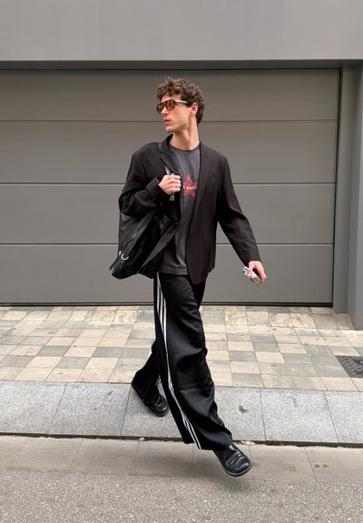 Joven caminando al aire libre con gafas de sol, blazer negro, camiseta gráfica, pantalones anchos negros con rayas blancas, zapatos negros y llevando una bolsa negra.
