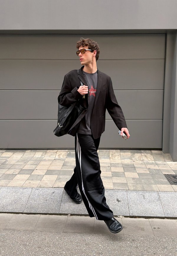 Jeune homme marchant dehors portant des lunettes de soleil, un blazer noir, un t-shirt graphique, un pantalon large noir avec des rayures blanches, des chaussures noires et portant un sac noir.
