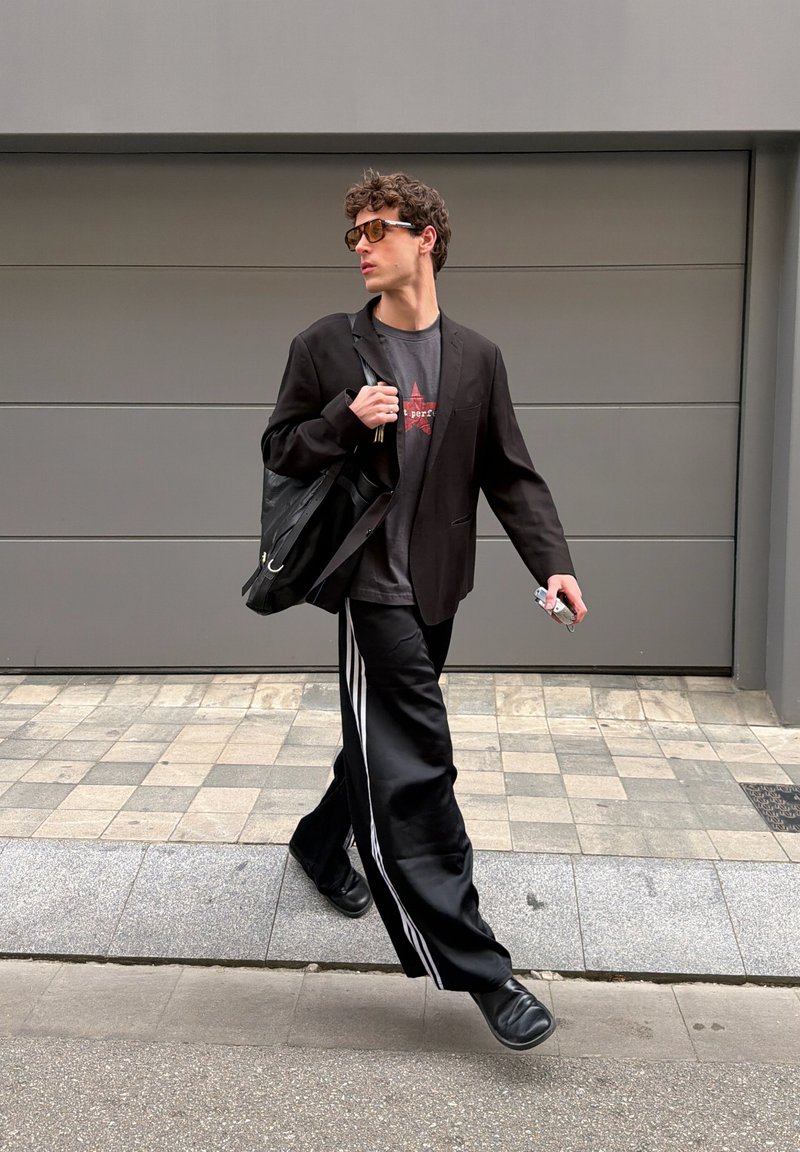 Jeune homme marchant dehors portant des lunettes de soleil, un blazer noir, un t-shirt graphique, un pantalon large noir avec des rayures blanches, des chaussures noires et portant un sac noir.