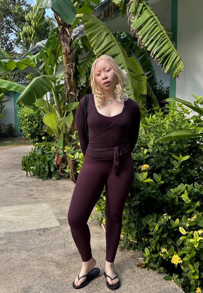 Top burdeos de manga larga con textura acanalada y detalle de lazo en la cintura, combinado con leggings a juego. Fondo de follaje verde exuberante y sendero.