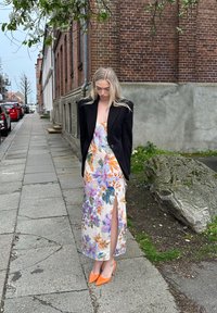 Blazer negro sobre un vestido crema con estampado floral y una abertura lateral. El conjunto se complementa con unos tacones de punta naranja brillante.