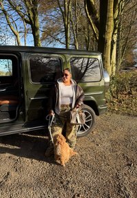 Grüner SUV auf Schotter geparkt. Person in brauner Jacke hält einen Hund an der Leine. Tarnhose und eine Handtasche mit Lederdetails.