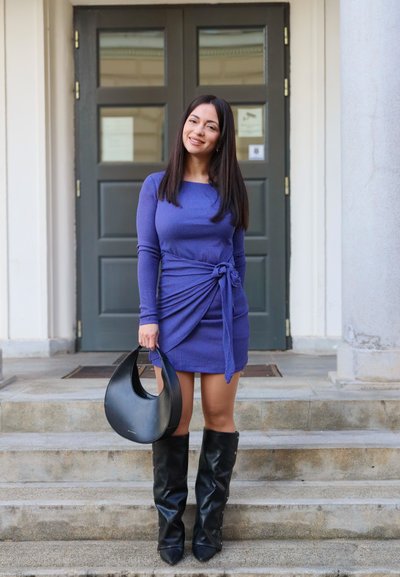 Mujer de pie en los escalones frente a unas puertas dobles oscuras, vistiendo un vestido azul, botas negras hasta la rodilla y sosteniendo un bolso negro.