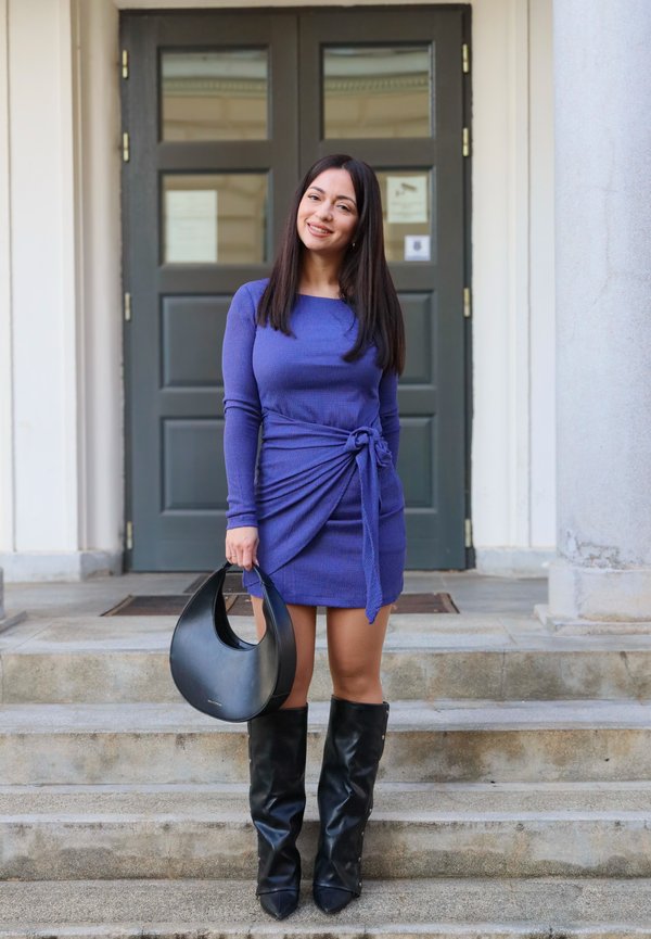 Kvinne som står på trappen foran mørke dobbeltdører, iført en blå kjole, svarte knelange støvler, og holder en svart håndveske.