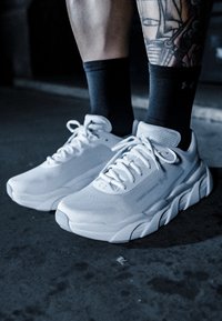 Žmogus, dėvintis baltus sportinius batelius ir juodas kojines, stovintis ant tamsios tekstūruotos dangos, matomas tatuiruotė vienoje kojoje.