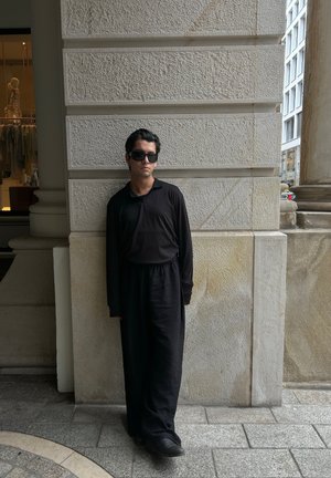 Top de manga larga de canalé negro y falda larga a juego, de pie contra una pared texturizada. Zapatos lisos y gafas de sol complementan el conjunto.