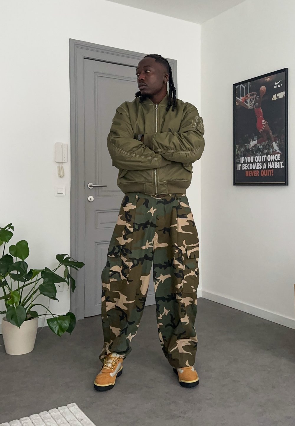 Blouson aviateur vert olive avec poignets côtelés, pantalon camouflage ample en vert et beige, et bottes jaunes. Intérieur avec une plante.