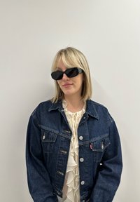 Μαύρα ορθογώνια γυαλιά ηλίου, τζην μπουφάν με κουμπιά και τσέπες, κρεμμώδης μπλούζα με φούσκες, ανοιχτό ξανθό μαλλί, ουδέτερο φόντο.