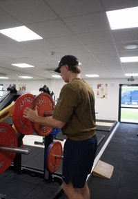 Máquina de levantamiento de pesas con discos de peso naranja y azul; una persona ajustando los pesos; gimnasio interior con iluminación brillante y espejos.