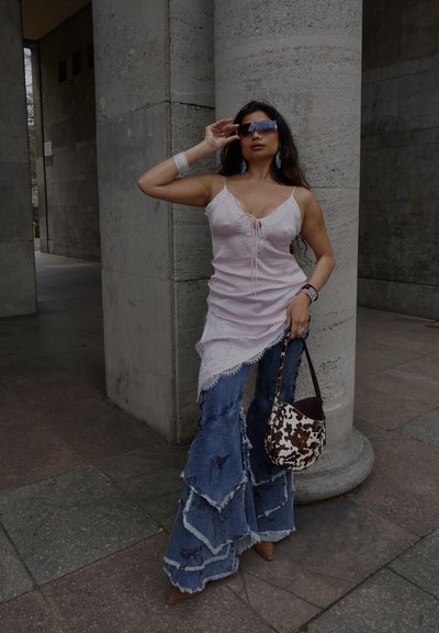 Mujer con un top asimétrico rosa, jeans anchos de varias capas, grandes gafas de sol, sosteniendo un bolso marrón y blanco con estampado, de pie junto a columnas de piedra.