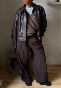 Άτομο που φοράει σκούρο καφέ δερμάτινο μπουφάν πάνω από καφέ πουλόβερ και παντελόνια με πλατύ πόδι, κρατώντας τσάντα σετ στο ίδιο σκούρο καφέ χρώμα, σε εσωτερικό χώρο.