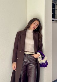 Γυναίκα που ακουμπά σε μια λευκή γωνιακή τοίχο, φοράει σκούρο καφέ παλτό, εκρού ριπ πουλόβερ, σκούρο δερμάτινο παντελόνι και κρατά μια μωβ τσάντα με φτερά.