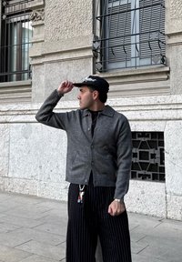 Uomo con cardigan grigio abbottonato e pantaloni neri a righe sottili che si sistema un berretto nero, in piedi vicino a un edificio in pietra con finestre con sbarre.