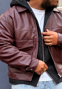 Veste en cuir marron avec une doublure en polaire noire, fermeture éclair et poche poitrine. Associée à un t-shirt blanc et un jean bleu.