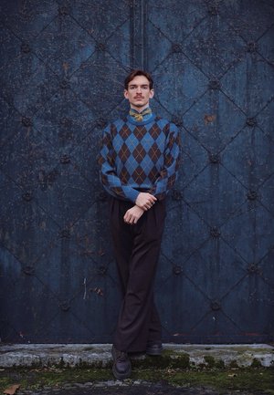 Hombre joven con bigote, vestido con suéter de argyle azul y pantalones negros, de pie con las piernas cruzadas frente a una puerta metálica azul texturizada.