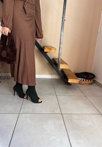 Καφέ, ριγέ μάξι φόρεμα με μακριά μανίκια, συνδυασμένο με μαύρες πλεκτές μπότες με ανοιχτό μπροστινό μέρος και τακούνι. Κρατάει μία καφέ τσάντα από ψεύτικη γούνα. Χαλικοστρωμένο γκρι πάτωμα.