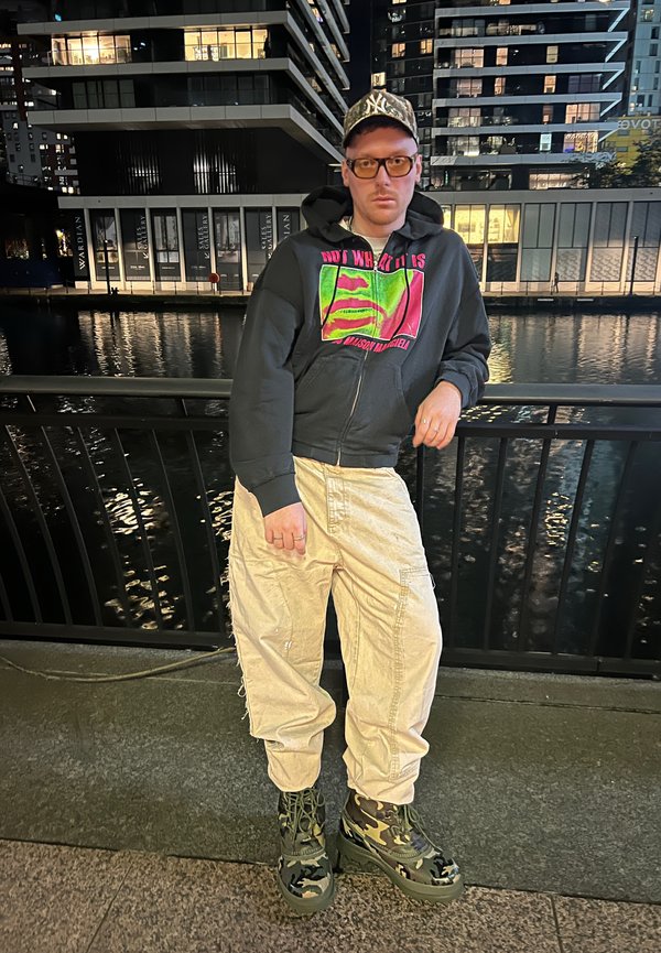 Sudadera con capucha y gráfico en neón, pantalones cargo crema y botas de plataforma de camuflaje. Escenario urbano nocturno con reflejos de agua en el fondo.